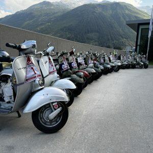 Mehrere Motorroller sind nebeneinander aufgereiht, im Hintergrund sind Berge und ein modernes Gebäude sichtbar.