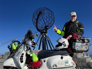 Olaf Just mit seiner beladenen weißen Vespa GTS 300 stehend vor dem Globe am Nordkapp bei strahlend sonnigem Wetter