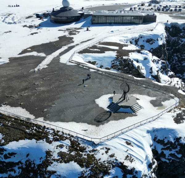 Blick aus der Luft (Drohnenaufnahme) des Nordkap-Globus - ringsrum teilweise Schnee und im Hintergrund das Gebäude des Informationscenter am Nordkap.