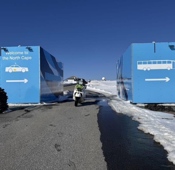 Ein Motorradfahrer fährt zwischen zwei großen blauen Containern auf einer verschneiten Straße mit klar blauem Himmel entlang.