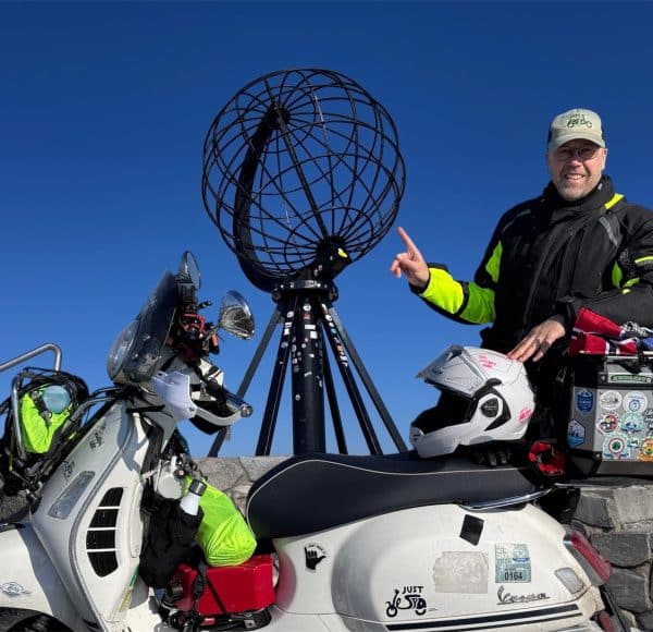 Vespa Fahrer Olaf Just mit schwarzer Jacke, grüner JustRide Cap und beladener weißer Vespa vor dem Nordkapp Globus unter strahlend blauem Himmel