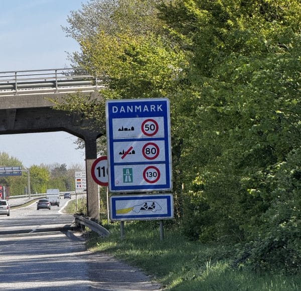 Verkehrsschild an der Grenze zu Dänemark mit Geschwindigkeitsbegrenzungen für verschiedene Straßenarten, blauem Hintergrund und Bäumen im Hintergrund.