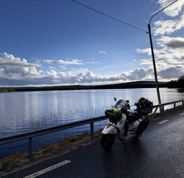 Bepackte weisse Vespa GTS steht auf einer nassen Straße östlich von Dorotea, Schweden neben einem See unter blauem Himmel mit Wolken, umgeben von Natur und einem Straßenlaterne. © JustKommunikation GmbH | JustRide it
