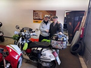 Zwei Personen stehen in der Werkstatt in Umea zwischen Motorrädern und der weißen Vespa GTS, lächeln und posieren für ein Foto. Im Hintergrund ein Schild mit der Aufschrift „Eagle Days“. © JustKommunikation GmbH | JustRide it