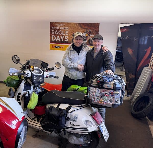 Zwei Personen stehen in der Werkstatt in Umea zwischen Motorrädern und der weißen Vespa GTS, lächeln und posieren für ein Foto. Im Hintergrund ein Schild mit der Aufschrift „Eagle Days“. © JustKommunikation GmbH | JustRide it