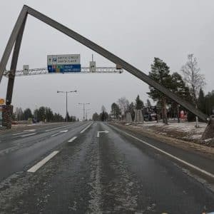 Blick auf das Santa Claus Werbekonstruktion über der Straße von der Fahrenden Vespa südlich von Rovanimie - rechts und links liegt Schnee und es sind Bäume sowie Büsche zu sehen.