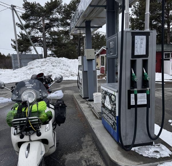 Bepackte weisse Vespa GTS steht an der Tanksäule einer Tankstelle nördlich von Inari / Finnland, im Hintergrund hohe Berge von Schnee am 6. Mai 2025.