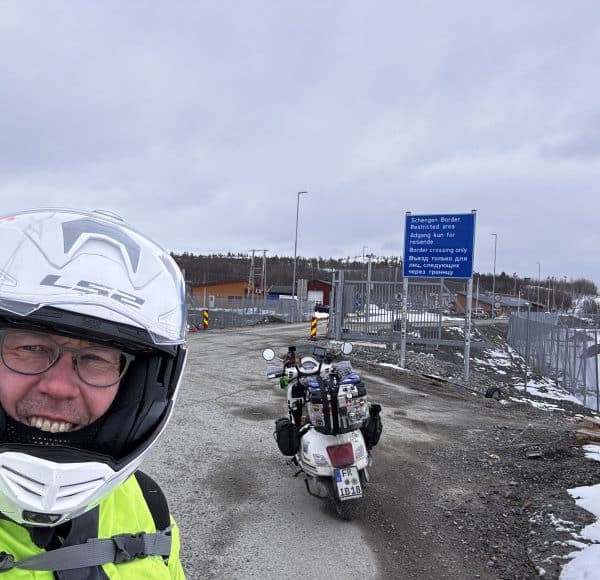 Motorradfahrer mit Helm lächelt, Motorrad auf grauer Straße, blauer Hinweisschild im Hintergrund, bewölkter Himmel, kalte Wetterbedingungen.