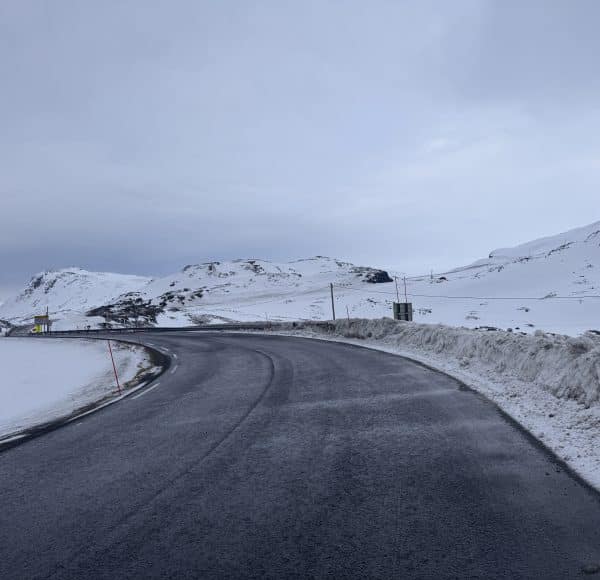 Verschneite Straße führt durch winterliche Hügellandschaft unter bewölktem Himmel.