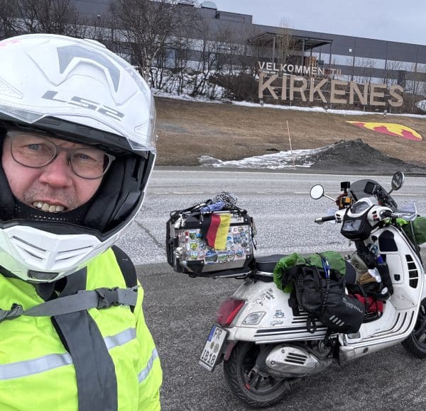 Person mit Motorradhelm macht Selfie vor einem Roller, im Hintergrund steht ein großes