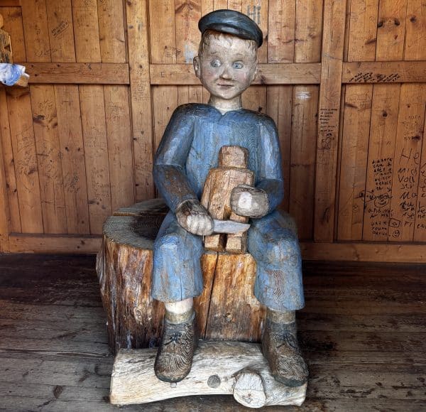 Holzskulptur eines Jungen mit Mütze, der auf einem Baumstamm sitzt und eine kleinere Holzfigur hält, vor Holzwand.