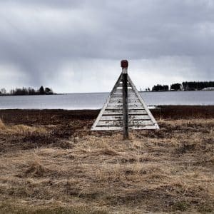 Dreieckiges hölzernes Seezeichen am Ufer, dahinter Wasser und grauer, bewölkter Himmel.