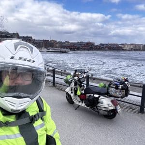 Person in Motorradkleidung mit Helm steht am Wasser, Motorrad im Hintergrund, bewölkter Himmel, Stadt im Hintergrund.