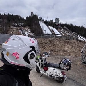 Person mit Motorradhelm steht vor einem Roller, im Hintergrund verschneite Skisprungschanzen und bewaldete Hügel unter bewölktem Himmel.