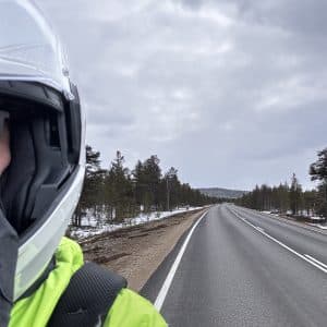 Motorradfahrer mit Helm und reflektierender Jacke auf leerer Landstraße, bewölkter Himmel, Bäume und leichte Schneereste am Straßenrand.
