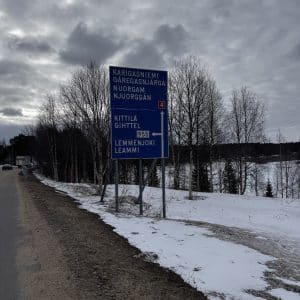 Verkehrsschild mit Wegweisern nach Kittilä und Lemmenjoki, steht an einer verschneiten Straße, umgeben von kahlen Bäumen unter bewölktem Himmel.