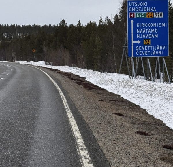 Eine Landstraße mit etwas Schnee am Rand, daneben ein blauer Wegweiser mit Ortsnamen auf Finnisch und Samisch, Wald im Hintergrund.