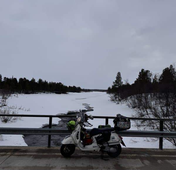 Ein weißer Motorroller steht auf einer Brücke, im Hintergrund ein verschneiter Fluss und bewaldete Landschaft unter grauem Himmel.