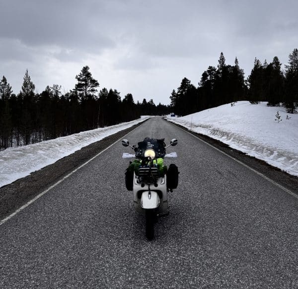 Motorrad steht auf einer leeren Straße, umgeben von Schnee und Bäumen unter bewölktem Himmel.