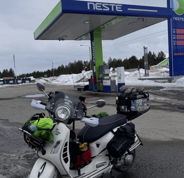 Roller steht an einer Tankstelle mit grün-blauem Dach, umgeben von etwas Schnee und bewölktem Himmel.