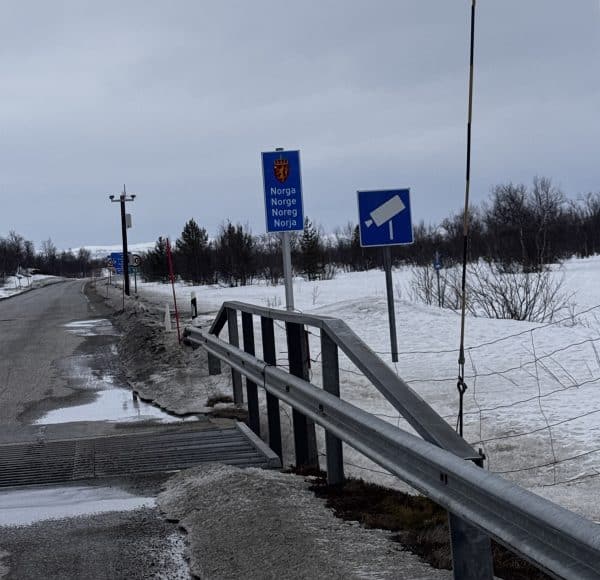 Verschneite Landstraße mit Leitplanke, Verkehrsschildern und kahlen Bäumen im Hintergrund unter bewölktem Himmel.