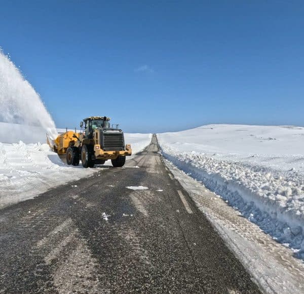 Ein Schneepflug räumt eine schneebedeckte Landstraße bei klarem, blauem Himmel.