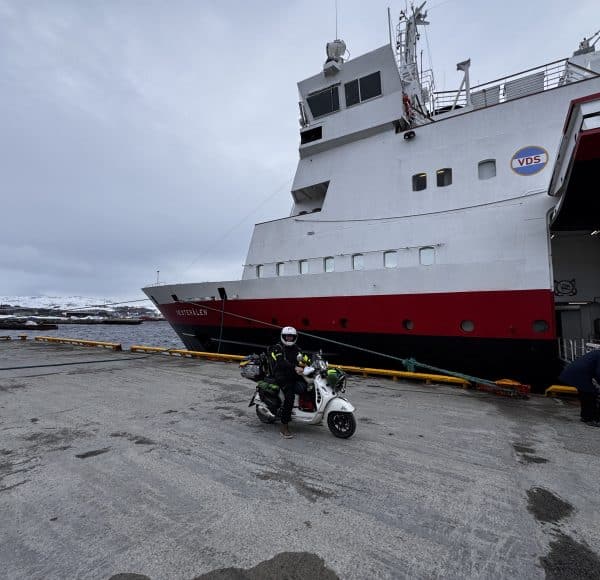 Person auf einem Roller steht vor einem großen weißen und roten Schiff im Hafen bei bewölktem Himmel.