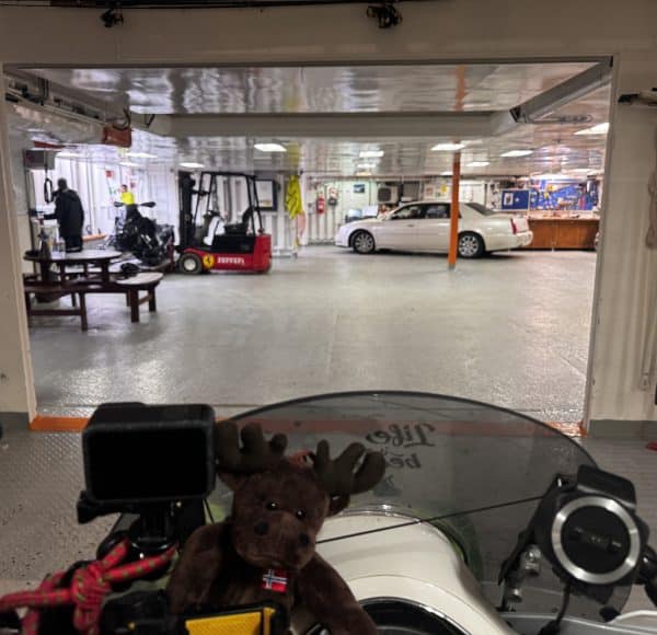 Innenraum einer Fähre mit geparkten Autos, Personen im Hintergrund und einem Motorrad mit Steuerrad und Plüschtier im Vordergrund.