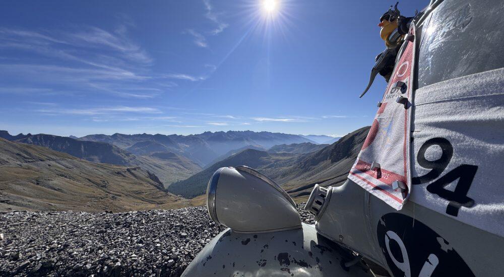 Hubschrauber auf Berghügel, Person schaut hinaus, sonniger Himmel, Gebirgslandschaft im Hintergrund.
