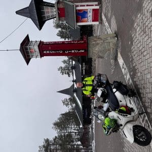 Person mit Motorrad vor einem roten Turm und Gebäuden, winterliche Umgebung, leicht bewölkter Himmel.
