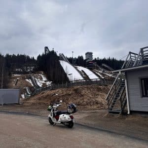 Eine verschneite Skisprungschanze mit einem weißen Motorroller im Vordergrund und wolkigem Himmel im Hintergrund.