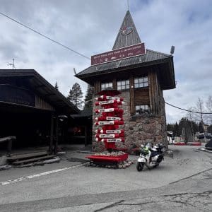 Holzgebäude mit spitzem Turm, daneben Straßenschild mit vielen Richtungen und ein geparktes Motorrad, bewölkter Himmel.