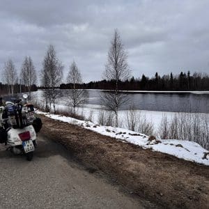 Motorrad am Straßenrand, Blick auf einen verschneiten Fluss und kahle Bäume unter bewölktem Himmel.