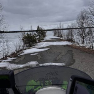Straße mit schmelzendem Schnee, Bäumen am Rand und bewölktem Himmel, gesehen aus der Perspektive eines Motorrads oder Fahrrads.