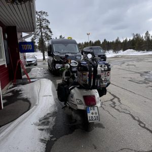 Ein Motorrad und ein Auto stehen auf einem winterlichen Parkplatz neben einem Gebäude, Schnee liegt am Rand, bewölkter Himmel.