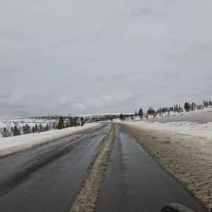 Nasse Straße mit Schnee an den Seiten, bewölkter Himmel, kahle Landschaft und einige Bäume im Hintergrund.