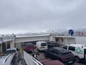 Mehrere Autos und ein Motorrad stehen auf einer Autofähre, im Hintergrund ist nebliges Wetter über dem Wasser zu sehen.