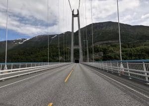 Leere Hängebrücke über einem Fluss, Berge im Hintergrund, bewölkter Himmel, Fahrbahn mittig fotografiert.