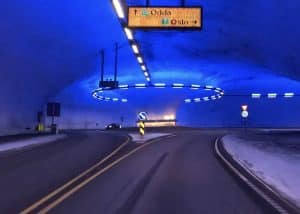 Straßentunnel mit blau beleuchtetem Innenraum und einem Kreisverkehr, Straßenschilder sichtbar.