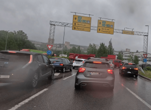 Autos stehen im Regen im dichten Verkehr auf einer mehrspurigen Straße, gelbe Richtungsschilder sind sichtbar.