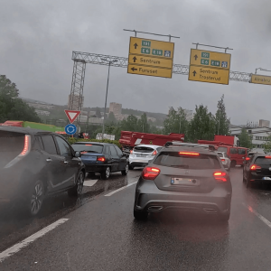 Autos stehen im Regen im dichten Verkehr auf einer mehrspurigen Straße, gelbe Richtungsschilder sind sichtbar.