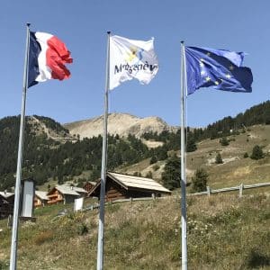 Drei Fahnen wehen im Wind vor Bergen und Holzhäusern: Frankreich, Argentière und Europäische Union.