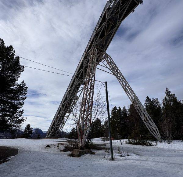 Große Metallbrücke über verschneite Landschaft, mit Bäumen im Hintergrund und bewölktem Himmel.