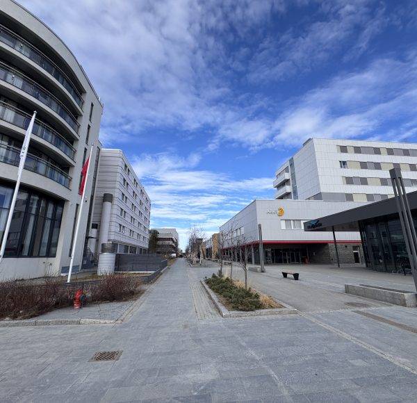 Moderne Gebäude mit Glasfassaden und weitem Gehweg, blauer Himmel mit Wolken, städtische Umgebung, keine Menschen sichtbar.