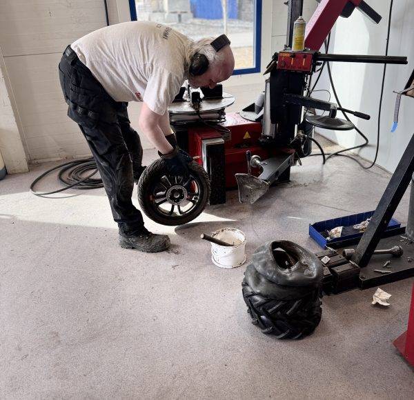 Ein Mechaniker arbeitet in einer Werkstatt und hantiert mit einem Autoreifen an einer Montagemaschine. Werkzeuge und weitere Reifen liegen in der Nähe.