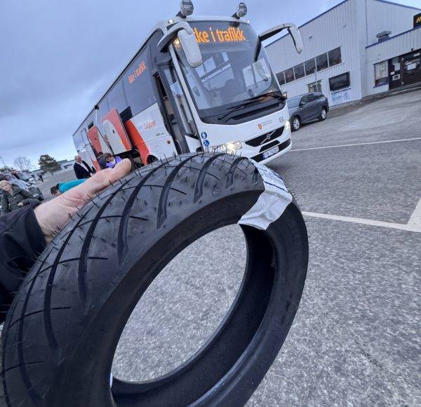 Nahaufnahme eines Reifens, der vor einem geparkten Bus auf einem Parkplatz mit Industriegebäuden im Hintergrund gehalten wird.