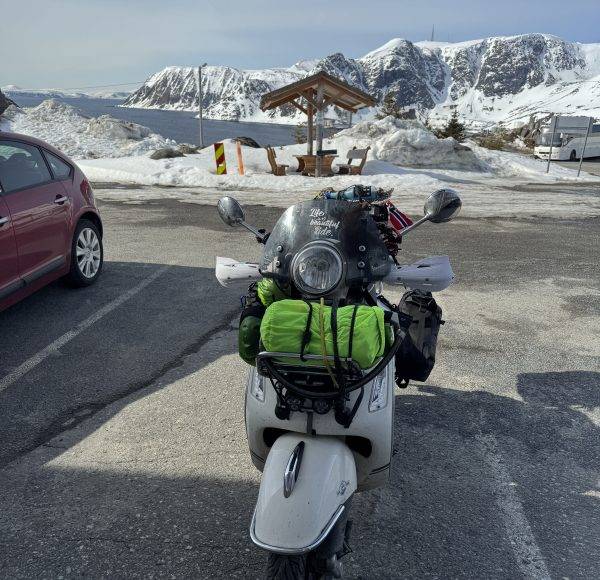 Ein Motorrad mit grüner Gepäcktasche steht auf einem verschneiten Parkplatz, Berge und ein Auto im Hintergrund.