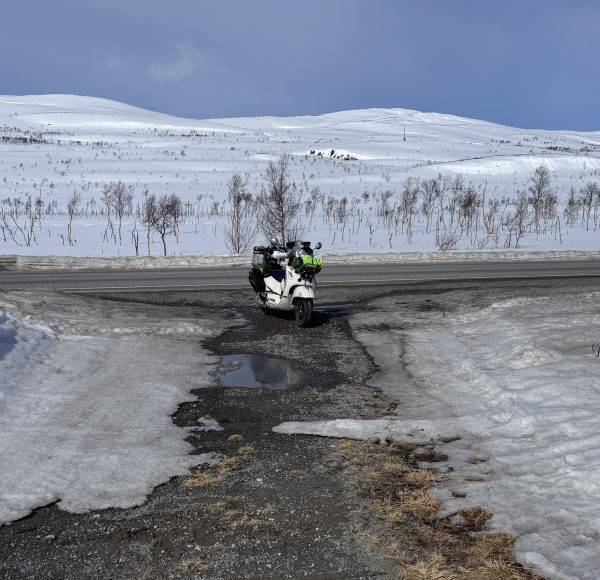 Motorrad fährt auf einer nassen Straße durch eine schneebedeckte Landschaft mit kahlen Bäumen und Hügeln im Hintergrund.