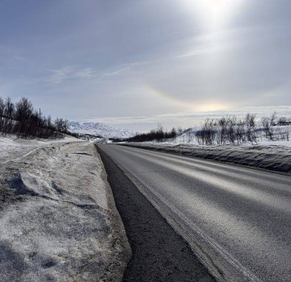 Verschneite Straße bei Sonnenlicht, Berge im Hintergrund, klare winterliche Landschaft.