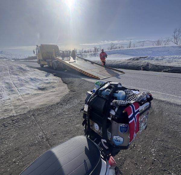 Verschneite Straße mit Motorrad vorne, norwegische Flagge sichtbar, Menschen in Winterkleidung laufen, Sonne scheint, klare Winterlandschaft.